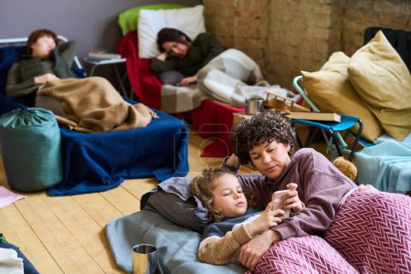 Téléchargez les photos : Garçon mignon avec smartphone et sa mère regardant l'écran tout en se reposant au lit ou en dormant contre deux autres réfugiées - en image libre de droit
