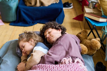 Foto de Adorable chico con el pelo rubio rizado y su madre durmiendo bajo cuadros de manta en el suelo de la habitación con colchones para refugiados - Imagen libre de derechos