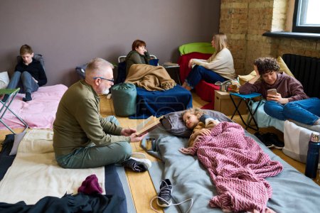 Téléchargez les photos : Homme mûr avec livre ouvert lecture conte de fées à son petit-fils couché dans son lit tout en se reposant dans un abri temporaire avec d'autres réfugiés - en image libre de droit