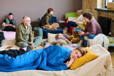 Téléchargez les photos : Adolescent garçon garder sa tête sur l'oreiller tout en dormant sous la couverture bleue contre le groupe de personnes communiquant sur leurs lits - en image libre de droit