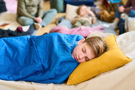 Photo for Peaceful schoolboy napping on sleeping place under blue blanket while keeping his head on yellow pillow against group of refugees - Royalty Free Image