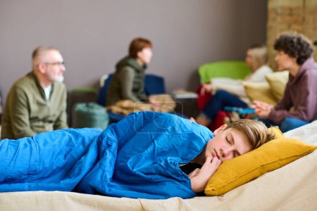 Photo for Cute serene boy with his head on pillow sleeping under blanket on couchette against roup of refugees discussing latest news - Royalty Free Image