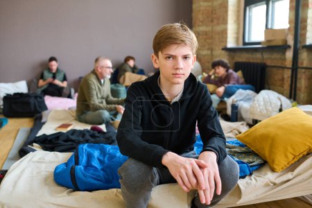 Téléchargez les photos : Garçon sérieux en tenue décontractée assis sur la couchette préparé pour les sans-abri et regardant la caméra contre les réfugiés parler entre eux - en image libre de droit
