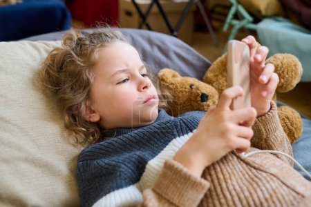 Photo for Cute blond boy in casual knitted sweater lying on couchette and scrolling in his mobile phone while having rest in temporary shelter - Royalty Free Image