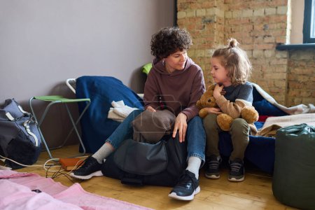 Foto de Mujer joven en ropa casual mirando a su hijo mientras está sentada en la sofá junto a él y sacando el suéter gris de lana caliente de la bolsa - Imagen libre de derechos