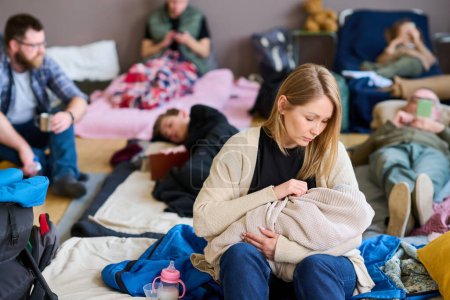 Junge blonde Flüchtlingsfrau wiegt Baby in warmem Plaid, während sie auf dem Bett sitzt, das von Freiwilligen für Menschen in Schwierigkeiten vorbereitet wird