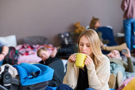 Téléchargez les photos : Young blond female refugee with yellow mug having hot tea while sitting on couchette by stroller with her baby against other people - en image libre de droit