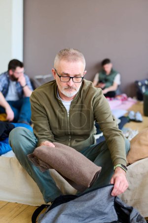 Foto de Hombre maduro en anteojos y ropa casual bolsa de embalaje mientras está sentado en la cama o sofá en el campo de refugiados para las personas necesitadas y problemas - Imagen libre de derechos