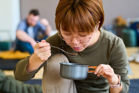Photo for Young Chinese female refugee eating soup or other self prepared food in refugee camp against other temporarily homeless people - Royalty Free Image