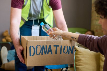 Téléchargez les photos : Jeune homme bénévole en uniforme tenant une boîte de dons avec de la nourriture et des boissons pendant que la réfugiée prend une bouteille d'eau - en image libre de droit