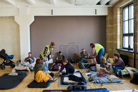 Foto de Dos jóvenes voluntarios difunden ayuda humanitaria entre refugiados sentados en sus lugares de dormir en el suelo de una amplia habitación - Imagen libre de derechos