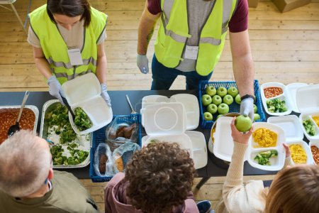 Téléchargez les photos : Au-dessus de l'angle de deux volontaires en uniforme répandant de la nourriture parmi les réfugiés avec des conteneurs debout dans la file d'attente par table - en image libre de droit