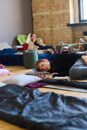 Téléchargez les photos : Écolier avec téléphone portable reposant sur la place de sommeil sur le sol et texter contre une jeune réfugiée couchée sur une couchette - en image libre de droit
