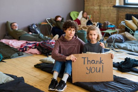 Téléchargez les photos : Jeune femme reconnaissante et son petit fils exprimant leur gratitude aux bénévoles assis sur le lieu de sommeil sur le sol du camp - en image libre de droit