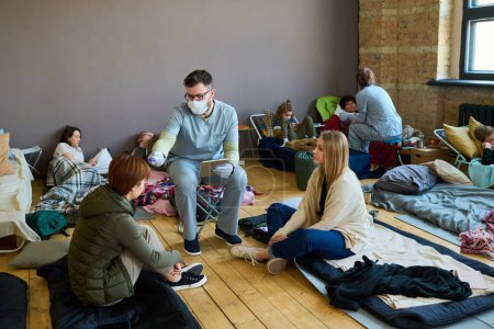 Männlicher Freiwilliger oder Gesundheitsarbeiter misst die Körpertemperatur einer jungen Flüchtlingsfrau, die auf ihrem Schlafplatz auf dem Fußboden sitzt