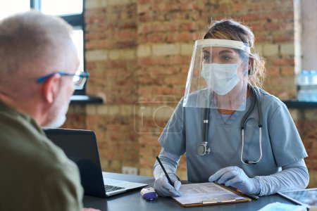 Téléchargez les photos : Jeune clinicienne dans un écran protecteur, un masque et un gommage médical à l'écoute d'un réfugié masculin mature décrivant ses symptômes - en image libre de droit