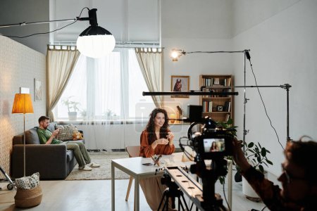Téléchargez les photos : Heureuse jeune blogueuse assise à table dans un loft pendant l'enregistrement vidéo d'une publicité filmée par un cameraman - en image libre de droit