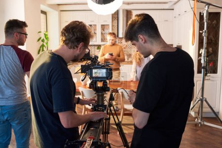 Téléchargez les photos : Rear view of videographer and his assistant adjusting video camera for shooting of commercial or cooking masterclass in the kitchen - en image libre de droit