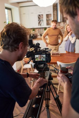 Photo for Two young videographers moving video camera along slider during shooting of commercial or cooking masterclass in the kitchen - Royalty Free Image
