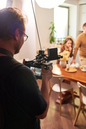 Téléchargez les photos : Jeune opérateur ou vidéaste avec vidéocamera tournage cuisson masterclass ou publicité de repas faits maison ou pâtisserie - en image libre de droit