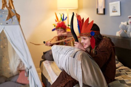 Foto de Happy cute girl in headdress of native Americans shooting from bow while sitting on bed during play with her friend or brother - Imagen libre de derechos