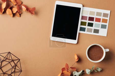 Téléchargez les photos : Top view of workplace of designer with tablet, color swatches, cup of coffee, handmade vase and dry orange leaves on beige table - en image libre de droit