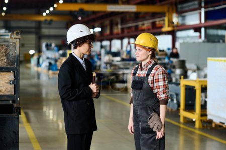 Foto de Young female engineer in workwear and inspection manager in black suit standing in large workshop of modern factory and communicating - Imagen libre de derechos