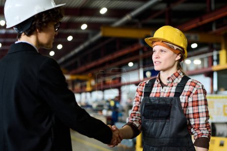 Téléchargez les photos : Young female engineer in workwear looking at factory manager during handshake while both standing in front of each other - en image libre de droit
