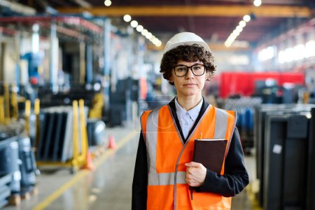 Photo for Young serious female factory manager in workwear and protective helmet looking at camera while standing in large workshop or warehouse - Royalty Free Image