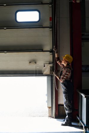 Foto de Young female engineer opening shutter roller door of garage, hangar or workshop while standing inside and pulling long chain down - Imagen libre de derechos