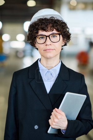 Photo for Young serious female manager of modern factory wearing hardhat and black suit holding tablet while looking at camera in warehouse - Royalty Free Image