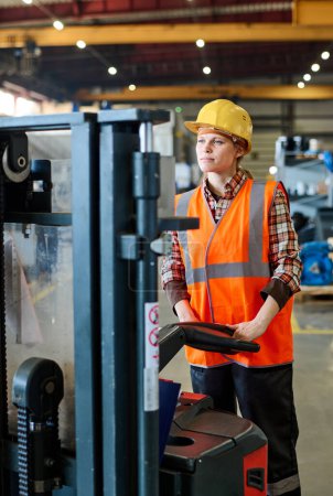 Téléchargez les photos : Female engineer looking forwards while standing in front of electric forklift and moving along aisle of workshop or warehouse - en image libre de droit