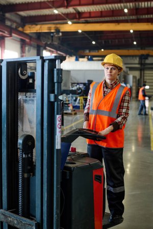 Foto de Young woman in hardhat, coveralls and orange jacket standing on forklift while moving along wide aisle of warehouse or workshop - Imagen libre de derechos