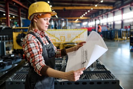 Téléchargez les photos : Side view of young female engineer in workwear holding unrolled blueprint with sketch of construction or machine part - en image libre de droit