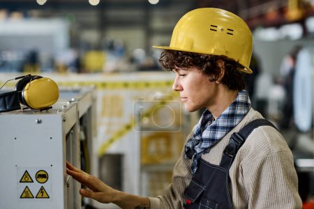 Téléchargez les photos : Young female engineer or technician in workwear and protective helmet checking industrial equipment or machine while working in factory - en image libre de droit