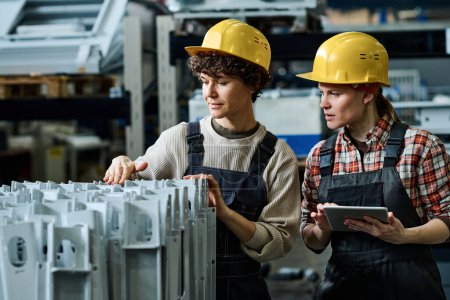 Téléchargez les photos : Jeune ingénieur avec tablette debout à côté d'un collègue vérifiant la qualité de nouveaux équipements ou de pièces de machines industrielles dans l'usine - en image libre de droit