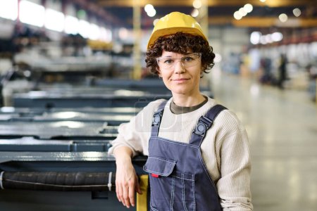 Foto de Young happy female worker of modern factory wearing grey sweater, blue coveralls and yellow hardhat standing by new industrial equipment - Imagen libre de derechos