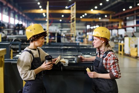 Photo for Two young engineers or workers of warehouse having sandwiches and hot tea at lunch break and discussing latest news or working points - Royalty Free Image