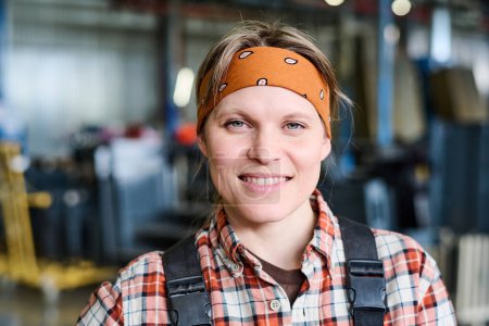 Photo for Happy young female engineer or factory worker in brown headband and checkered shirt looking at camera while standing in workshop - Royalty Free Image