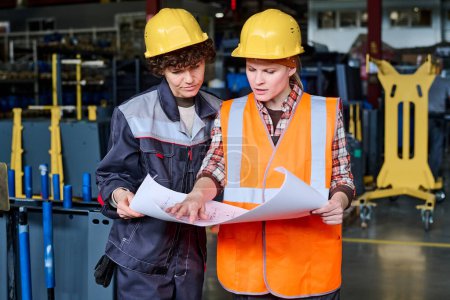 Foto de Dos trabajadores de la fábrica moderna que usan cascos de protección y ropa de trabajo mirando boceto en la impresión azul, mientras que uno de ellos explicándolo - Imagen libre de derechos
