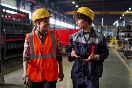 Photo for Two young female workers of modern factory discussing working points while moving down large workshop or distribution warehouse - Royalty Free Image