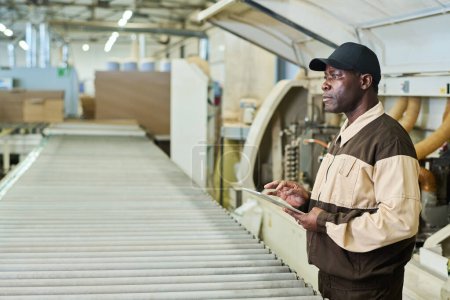 Téléchargez les photos : African foreman in uniform controlling machine with digital tablet during his work at factory - en image libre de droit