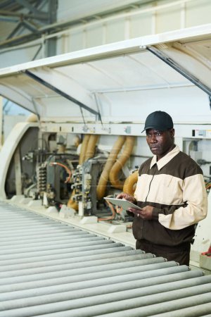 Téléchargez les photos : African serious worker in uniform using digital tablet during his work with machine at factory - en image libre de droit