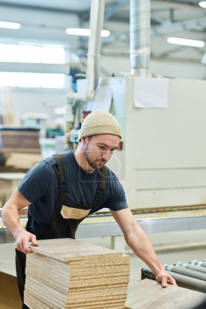 Téléchargez les photos : Jeune menuisier en uniforme travaillant avec des planches en bois à la machine à l'usine de meubles - en image libre de droit
