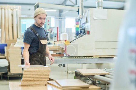 Téléchargez les photos : Jeune homme dans l'ensemble travaillant avec du bois à l'usine debout à la machine - en image libre de droit