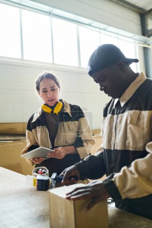 Téléchargez les photos : Travailleur africain en uniforme parlant à son collègue tout en emballant la boîte à table pour une exportation ultérieure - en image libre de droit