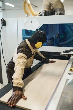 Téléchargez les photos : Travailleur africain dans un casque protecteur mettant de la planche de bois sur une machine à scier pour fabriquer des meubles à l'usine - en image libre de droit