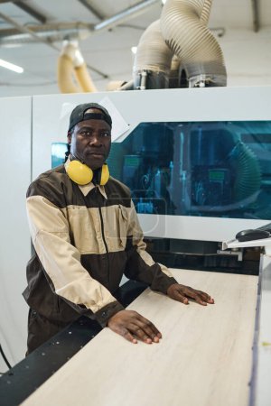 Foto de Portrait of African manual worker in uniform looking at camera while standing at modern equipment and working with wooden planks - Imagen libre de derechos