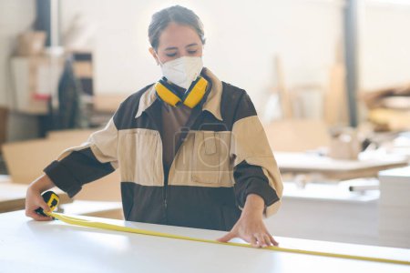 Foto de Trabajadora en máscara protectora usando cinta métrica para hacer mediciones en pieza de madera en la mesa - Imagen libre de derechos
