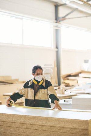 Foto de Mujer joven en máscara protectora y uniforme comprobando la longitud de los tablones de madera con cinta métrica - Imagen libre de derechos